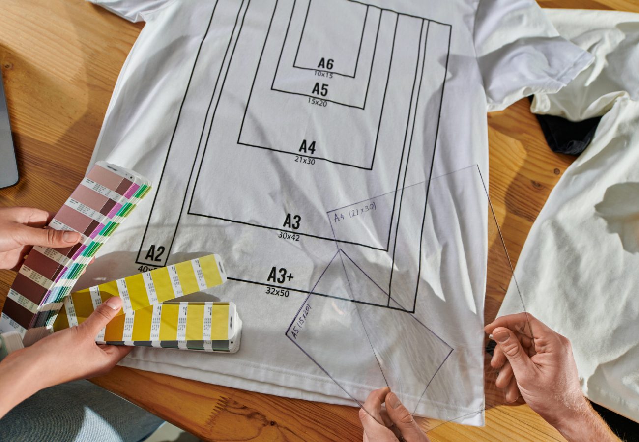 Top view of young multiethnic designers holding printing layers and color swatches while working near clothes with markup on table in print studio, thriving small enterprise concept Top view of young multiethnic designers holding printing layers and color swatches while working near clothes with markup on table in print studio, thriving small enterprise concept